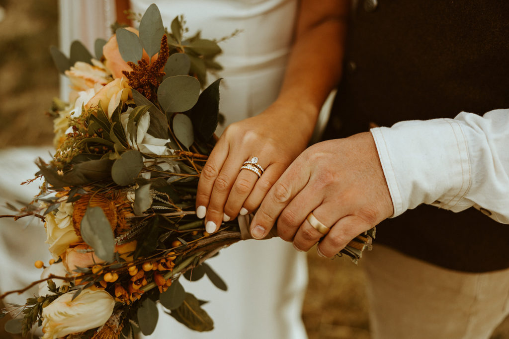 close up photo of bride and grooms hands holding fall wedding bouquet and rings